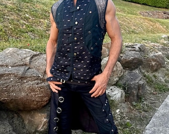 May include: A sleeveless black vest with decorative stitching and a high collar. The vest is paired with black pants featuring metal ring accents and lace-up details. The outfit is completed with black boots. The background features a natural outdoor setting with rocks and greenery.