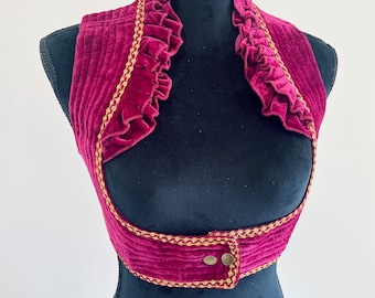May include: A burgundy velvet vest with gold trim and a ruffled collar. The vest has a button closure and is sleeveless.