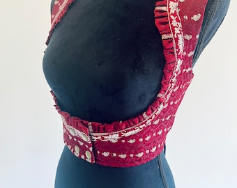 May include: A burgundy vest with silver floral embroidery and a ruffled trim. The vest features a button closure and is displayed on a black mannequin. The fabric has a subtle pattern of small, silver dots.