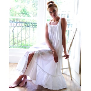 May include: A white cotton nightdress with a square neckline and a button-down front. The long, flowing nightdress features a ruffled hem and delicate lace trim. The woman is sitting on a white chair near a window.