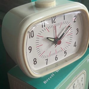 May include: Cream-coloured retro alarm clock with a rectangular face and rounded edges. The clock face has black numerals, red minute markers, and black hands with a red centre. The clock sits on a teal box.