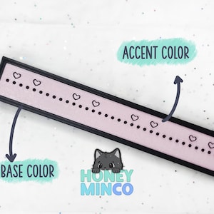 May include: A rectangular, black-framed decorative piece with a light pink center. The pink section features a dotted line with small heart accents. The text "HONEY MINCO" is visible in the lower right corner.