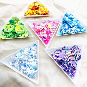 May include: A selection of colourful sequins displayed in white triangular dishes. The sequins are in shades of gold, green, pink, blue, and purple. Various shapes are present, including circles and flowers. Suitable for crafting and embellishments.