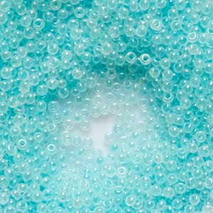 May include: A close-up image of a large quantity of small, light blue, round glass beads. The beads are arranged in a circular pattern, creating a gradient effect from a darker blue to a lighter blue.