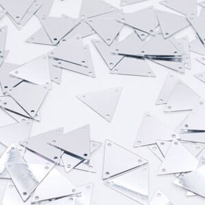 May include: A pile of silver triangle-shaped sequins with three holes in each. The sequins are scattered on a white background.
