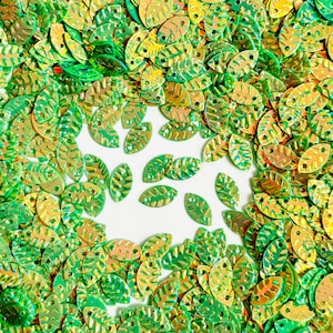 May include: A scattering of leaf-shaped sequins in green and gold hues. The sequins are spread across a white surface, creating a textured and decorative effect. Suitable for craft projects and embellishments.