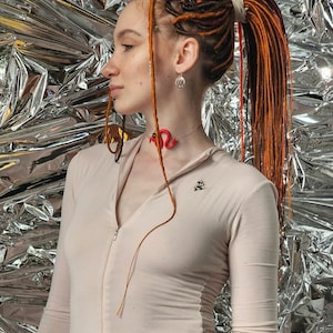 May include: A woman with long, red and brown dreadlocks styled in a ponytail. She is wearing a light brown, long-sleeved, zippered jumpsuit with a small embroidered design on the chest. The background is a silver, crinkled foil.