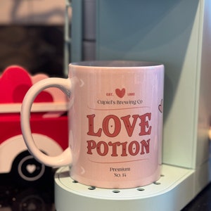 May include: A pink ceramic mug with a white handle. The mug has a pink and red design with the text "EST. 1892 Cupid's Brewing Co. LOVE POTION Premium No. 14".