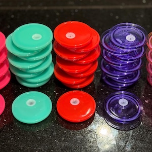 May include: Stacks of colorful plastic lids in pink, red, purple, turquoise and clear. The lids are round and have a slight dome shape.