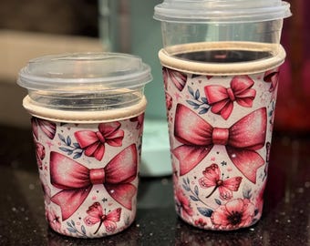 May include: Two reusable coffee cup sleeves with lids. The sleeves are decorated with a pattern of pink bows, butterflies, and flowers on a white background. The cups are designed to fit standard coffee cup sizes.