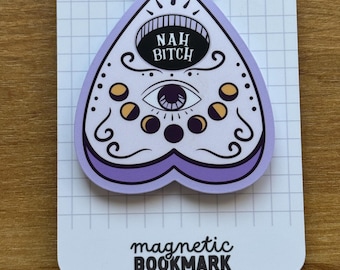May include: A magnetic bookmark with a purple and white planchette design. The planchette has an eye and moon phases, with the text "NAH BITCH" in a black oval. The bottom of the bookmark reads "magnetic BOOKMARK".