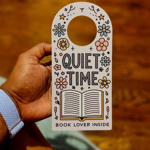 May include: White door hanger with a floral design and the words "QUIET TIME" and "BOOK LOVER INSIDE". The design includes an open book illustration and various flowers in pastel colors. The hanger has a circular cutout for the doorknob.