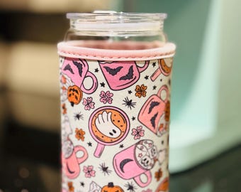 May include: A clear glass tumbler with a light pink fabric sleeve featuring a repeating pattern of pink mugs filled with ghost-shaped foam, orange jack-o'-lanterns, black bats, small flowers, and stars on a white background with a clear lid.