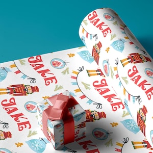 May include: White wrapping paper with a repeating pattern of nutcracker soldiers, gingerbread men, and the name "JAKE" in red and black lettering. A red and white gift box is wrapped in the paper.