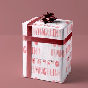 Puede incluir: Una caja de regalo blanca con una cinta y un lazo rojos. La caja está envuelta en papel con un patrón rosa y blanco de ropa de bebé y el nombre "Angelina" repetido.