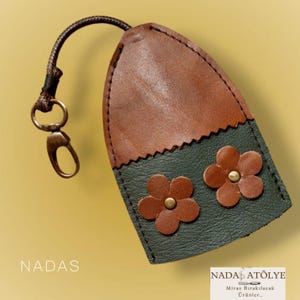 May include: A handcrafted leather key holder in brown and dark green, featuring two flower appliqués with brass studs. The key holder has a brown cord and a metal clasp. The word "NADAS" is printed on the bottom left.