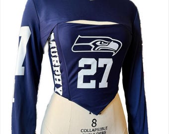 May include: A navy blue long-sleeved shirt with a white Seattle Seahawks logo and the number 27. The shirt has a cut-out at the chest and a pointed hem.