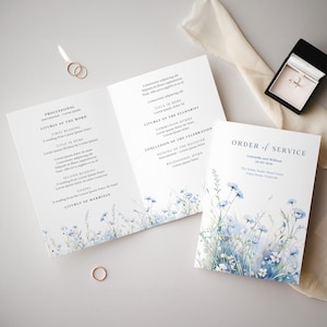 Könnte beinhalten: Ein Programmheft für eine Hochzeitszeremonie mit einem Aquarelldesign aus blauen Blumen und grünem Laub. Der Text auf der Vorderseite lautet "Ordnung des Gottesdienstes" mit den Namen "Gabrielle und William" und dem Datum "20.09.2020".