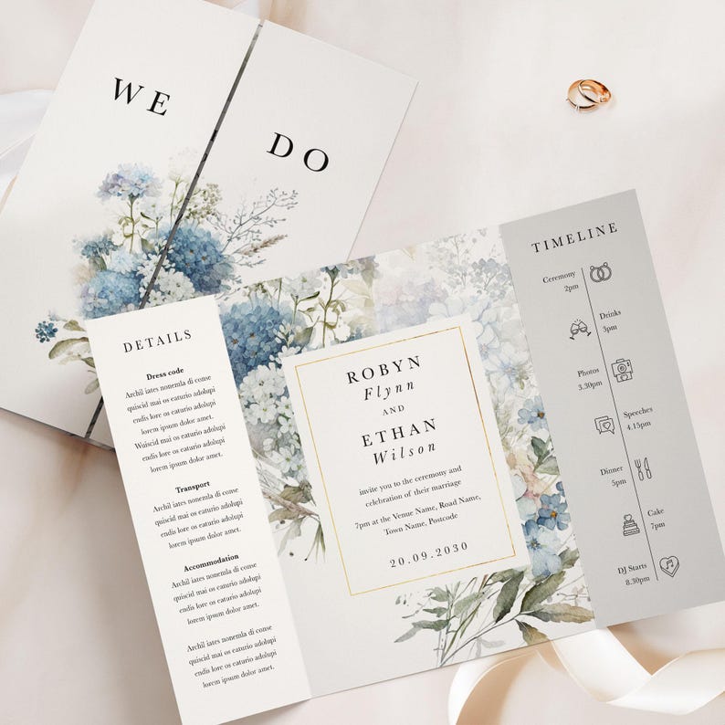 May include: Elegant wedding invitation suite featuring floral watercolor designs in shades of blue and white. The suite includes a main invitation, details card, and timeline card. Gold wedding rings are visible in the background.