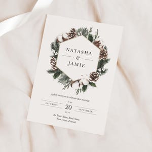 Op de afbeelding: Een huwelijksuitnodiging met een winterthema. De uitnodiging heeft een zeshoekig frame met een gouden rand, omringd door dennentakken, katoenen bollen en dennenappels. De tekst luidt "Natasha & Jamie" en bevat de datum en tijd van de bruiloft.