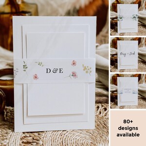 Op de afbeelding: Witte trouwuitnodigingssuite met gelaagd karton. Een bloemenband met de letters "D & E" wikkelt zich om het midden. Extra uitnodigingsontwerpen worden getoond, samen met de tekst "80+ ontwerpen beschikbaar."
