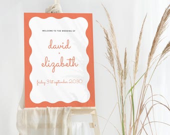 May include: A coral-colored wedding welcome sign with a scalloped white border. The sign reads "Welcome to the Wedding of David + Elizabeth" and the date "friday 31st september 2030" in a handwritten style font. The sign is displayed on a wooden easel.