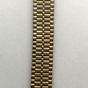 May include: A gold-tone metal watch band featuring a multi-link design. At the top, a rectangular plaque displays a small star emblem, two dark red circular stones, and the text Highland Park Cafeterias printed in a clear, sans-serif font.