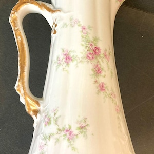 Op de afbeelding: Witte porseleinen kan met een gouden handvat en rand, versierd met roze bloemmotieven en groene bladeren. De kan heeft een uitlopende basis en een decoratieve tuit, met een elegant, vintage ontwerp.