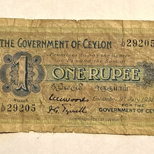 Puede incluir: Un billete vintage de una rupia del Gobierno de Ceilán, fechado el 9 de julio de 1931. El billete presenta un gran número "1" en azul y un borde decorativo. El texto "THE GOVERNMENT OF CEYLON" y "ONE RUPEE" está impreso en negro.
