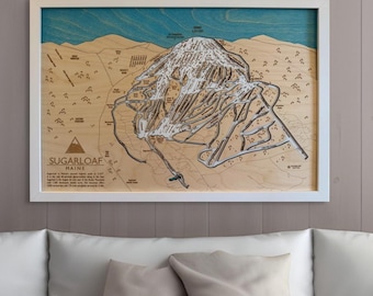 May include: A wooden map of Sugarloaf Mountain in Maine, showing the ski trails and lifts. The map is framed in white and has a blue and white background.