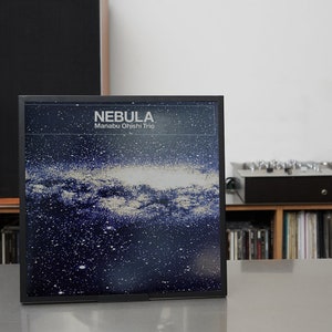 Könnte beinhalten: Ein gerahmtes Albumcover für "Nebula" von Manabu Ohishi Trio. Das Kunstwerk zeigt ein dunkelblaues und weißes Galaxiedesign. Der Albumtitel und der Künstlername stehen in weißer Schrift oben. Das Album wird auf einer hellen Oberfläche präsentiert.