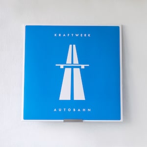 Puede incluir: Una portada de álbum cuadrada azul con el texto "KRAFTWERK" en blanco en la parte superior y "AUTOBAHN" en blanco en la parte inferior. Una imagen estilizada blanca de una autopista de dos carriles se encuentra en el centro de la portada.