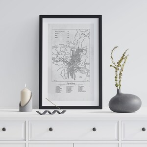 Pode incluir: Uma impressão emoldurada em preto e branco de um mapa intitulado "Plan of Meere" está pendurada acima de uma cômoda branca. Um vaso cinza com vegetação, uma vela em um suporte de concreto e um porta-incenso estão na cômoda. O mapa mostra um plano detalhado da cidade.