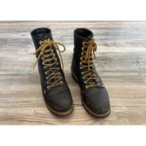 Puede incluir: Un par de botas de trabajo de cuero negro con diseño de caña alta, punteras redondeadas y ojales metálicos dorados. Las botas se ajustan con cordones gruesos de patrón amarillo y marrón y tienen una entresuela de color beige visible.