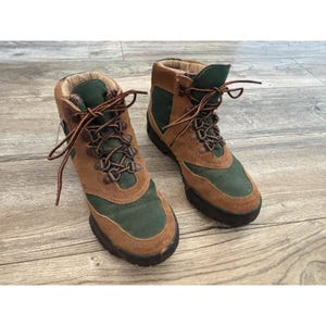 May include: A pair of ankle-high hiking boots featuring a combination of brown suede leather and dark green canvas panels. The boots include brown laces, metal eyelets, and thick black rubber soles with a textured tread for traction.