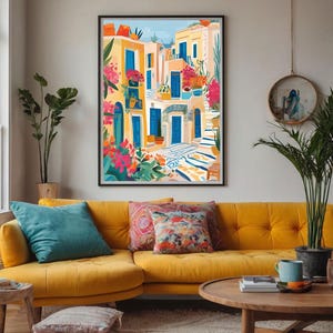 May include: A colorful illustration of a Mediterranean village with blue doors, yellow buildings, and lush green plants. The artwork is framed and hanging on a wall above a yellow couch with pillows.