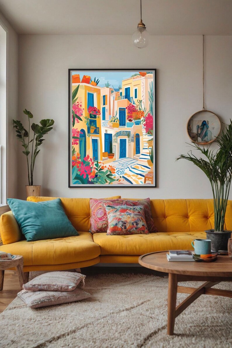 May include: A colorful illustration of a Mediterranean village with blue doors, yellow buildings, and lush green plants. The artwork is framed and hanging on a wall above a yellow couch with pillows.