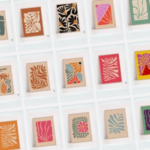 May include: A collection of framed art prints featuring abstract floral designs in various colors. The prints showcase botanical shapes in shades of orange, black, pink, and blue, set against neutral backgrounds. The art pieces are displayed in a grid.