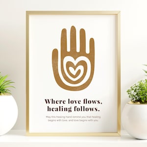 以下が含まれることがあります： 金色のフレームに入ったプリントで、茶色の手のデザインと中心にハートが描かれています。「Where love flows, healing follows」というテキストが下にあります。このプリントは、鉢植えの植物が置かれた白い棚に飾られています。