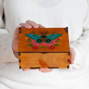 May include: A wooden box with a hinged lid, featuring a hand-painted design of a teal and black butterfly with pink and red floral accents. The box is a warm brown colour.