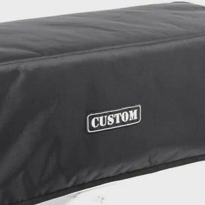 May include: A black rectangular protective cover made of a smooth, durable material. The cover features a centred, rectangular black patch with a white border and the word CUSTOM printed in bold, white, capitalised sans-serif lettering.