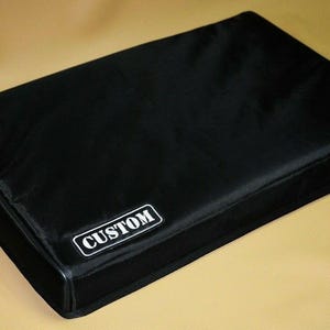 Può includere: Una copertura rettangolare nera imbottita con la parola "CUSTOM" in un rettangolo bianco. La copertura è realizzata in un materiale liscio, forse nylon. È probabilmente una custodia protettiva per apparecchiature.