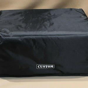 Puede incluir: Funda rectangular negra con la palabra "CUSTOM" en un rectángulo blanco. La funda parece estar hecha de un material duradero, posiblemente resistente al agua. La funda está sobre una superficie de color neutro.