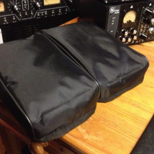 Puede incluir: Dos estuches negros rectangulares acolchados para equipos, probablemente de audio, sobre una superficie de madera. Los estuches tienen una textura lisa y ligeramente reflectante. Se ve equipo de audio en el fondo.