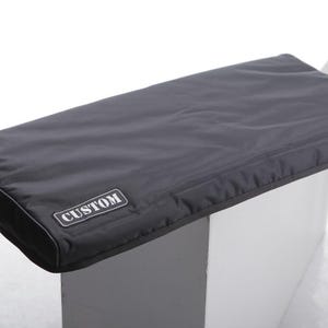 Op de afbeelding: Een zwarte, rechthoekige toetsenbordhoes met een gestikte rand en het woord "CUSTOM" op een wit label. De hoes is gemaakt van een glad, mogelijk waterbestendig materiaal en ligt op een wit oppervlak.