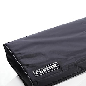 May include: A rectangular black protective case with a smooth, water-resistant fabric exterior. The case features a black rectangular patch with the white text CUSTOM stitched onto the front corner, along with black piping along the edges.