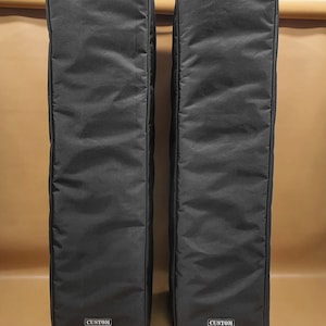 Puede incluir: Dos fundas negras y rectangulares para altavoces con una textura acolchada. Cada funda tiene una pequeña etiqueta rectangular en la parte inferior con la palabra "CUSTOM" en blanco. Las fundas están de pie sobre un fondo neutro.