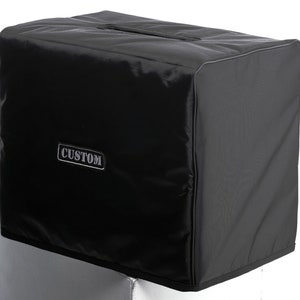 May include: A black rectangular cover with a handle on the top. The cover has a shiny, textured finish and a silver label that reads "CUSTOM". The cover is designed to fit over a rectangular object, possibly an amplifier or speaker.
