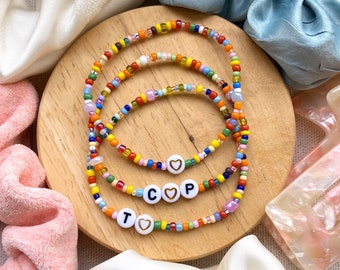 May include: Three colorful beaded bracelets with white letter beads spelling out "T", "C", and "P", each with a heart charm. The bracelets are on a wooden surface, surrounded by scrunchies and a hair clip.