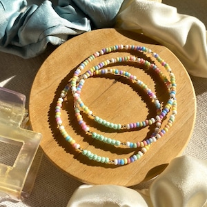 Könnte beinhalten: Drei pastellfarbene Perlenarmbänder mit goldenen Akzenten werden auf einer runden Holzoberfläche präsentiert. Die Armbänder weisen eine Mischung aus sanften Farben auf, darunter gelbe, rosa, blaue und grüne Perlen. Haarschmuck und Scrunchies im Hintergrund.